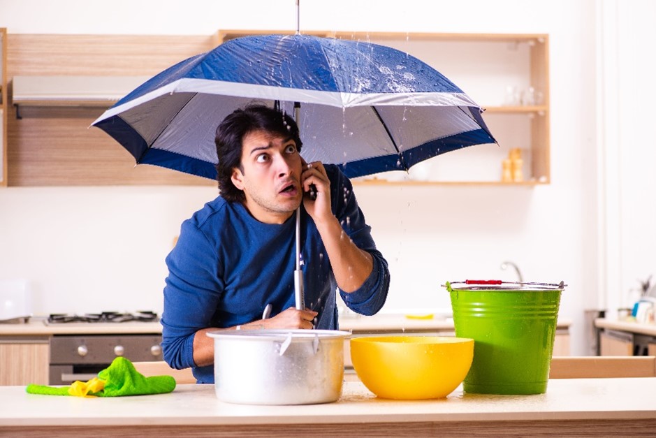 A male homeowner holds an umbrella in his kitchen to protect himself from a significant leak in the ceiling from a burst pipe. This qualifies as a plumbing emergency in Carol Stream, IL.